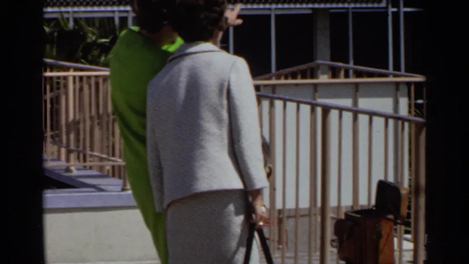 Two adult women in 1968 Scottsdale airport footage stand near metal railings, with luggage visible at the walkway edge.