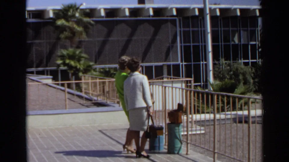 The women continue across the Scottsdale airport walkway, with luggage, railings, palms, and a modern terminal facade visible.