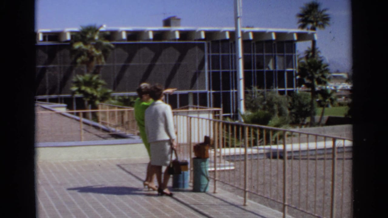 Two women stand with luggage on a tiled walkway near railings, palm trees, and a modernist building.