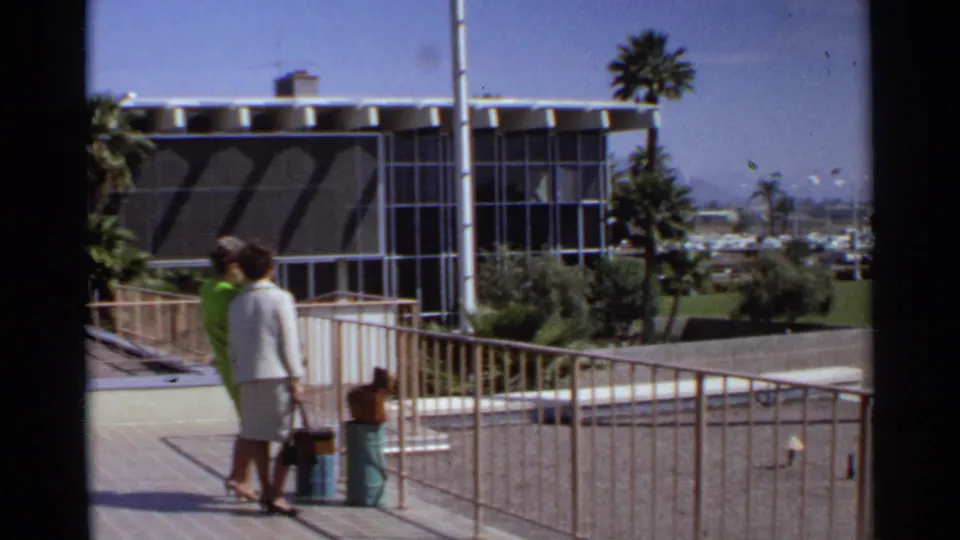 Two women in 1960s attire stand near luggage on a Scottsdale outdoor walkway, with palm trees and modernist architecture behind them.