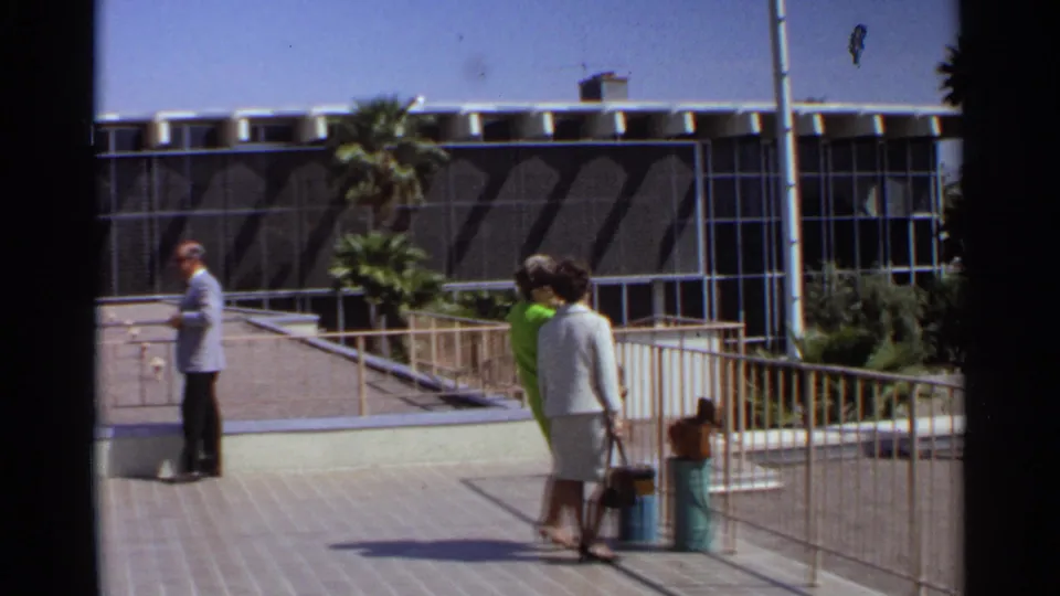 A suited man appears at the edge of the frame as two women with luggage stand on a Scottsdale walkway near palms and a modernist building.