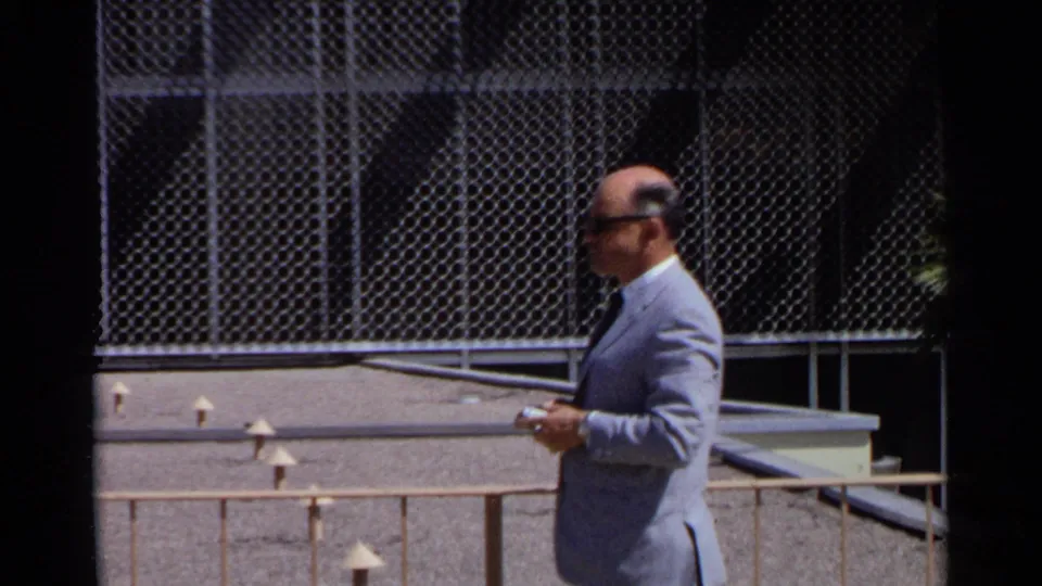 A man in a light suit and glasses passes a chain-link fence near the modernist building in the 1968 Scottsdale home-movie scene.