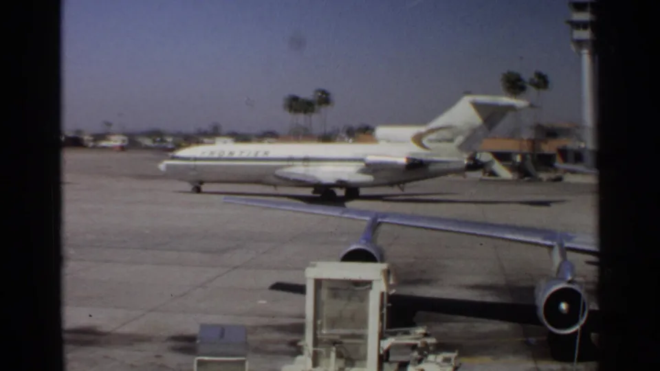 A Frontier-marked aircraft sits or rolls on the Scottsdale airport apron, framed by a terminal window and palm-lined background.
