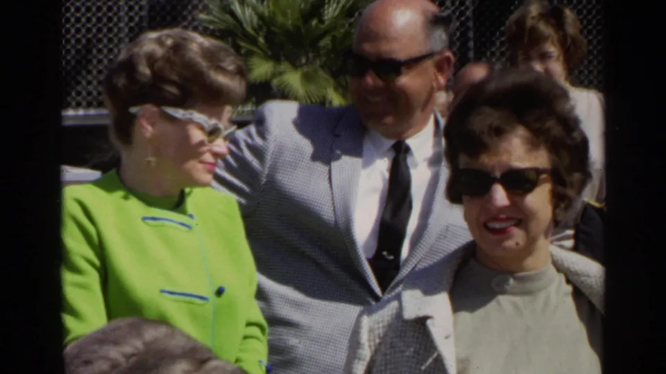 At Scottsdale Airport in 1968, three adult travelers smile near a fence, including a woman in a bright green jacket and white cat-eye sunglasses.