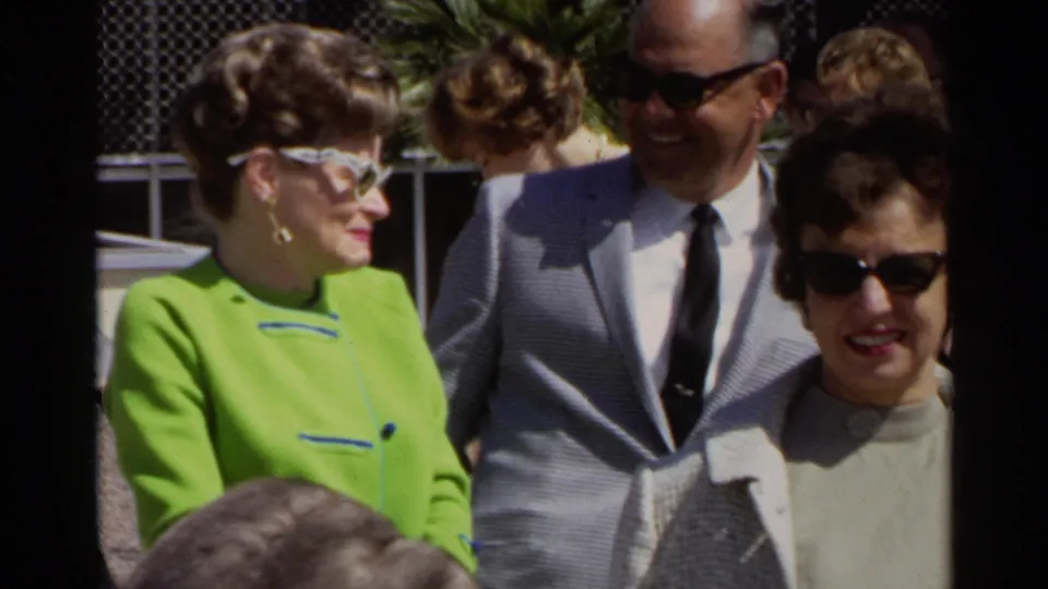 Three adult travelers in 1960s attire stand near the Scottsdale airport fence, smiling in bright sunlight.