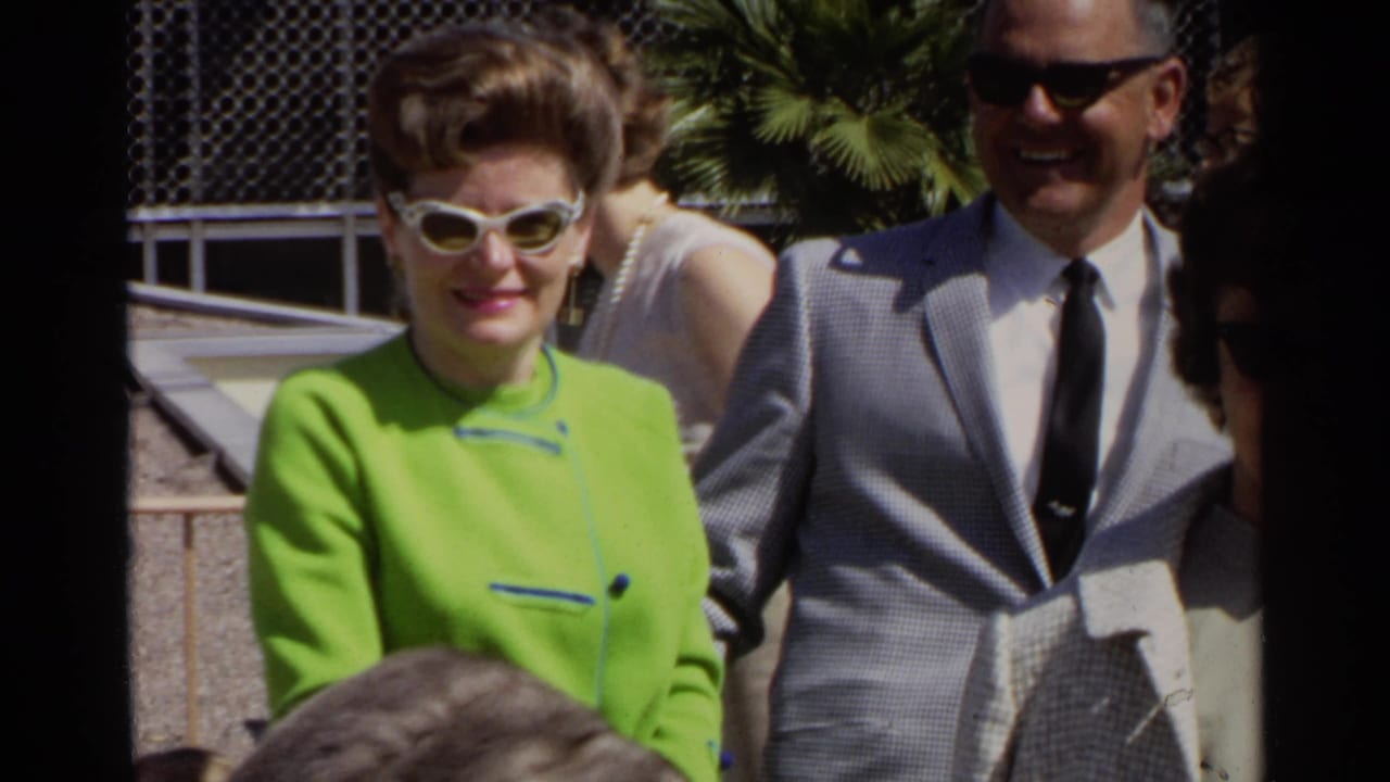 Woman in lime green outfit and white sunglasses standing beside a man in a gray suit outdoors.