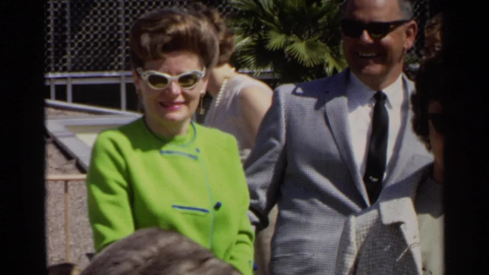 In 1968 Scottsdale footage, a woman in lime green and white cat-eye sunglasses stands near a smiling man in a gray suit at an outdoor event.