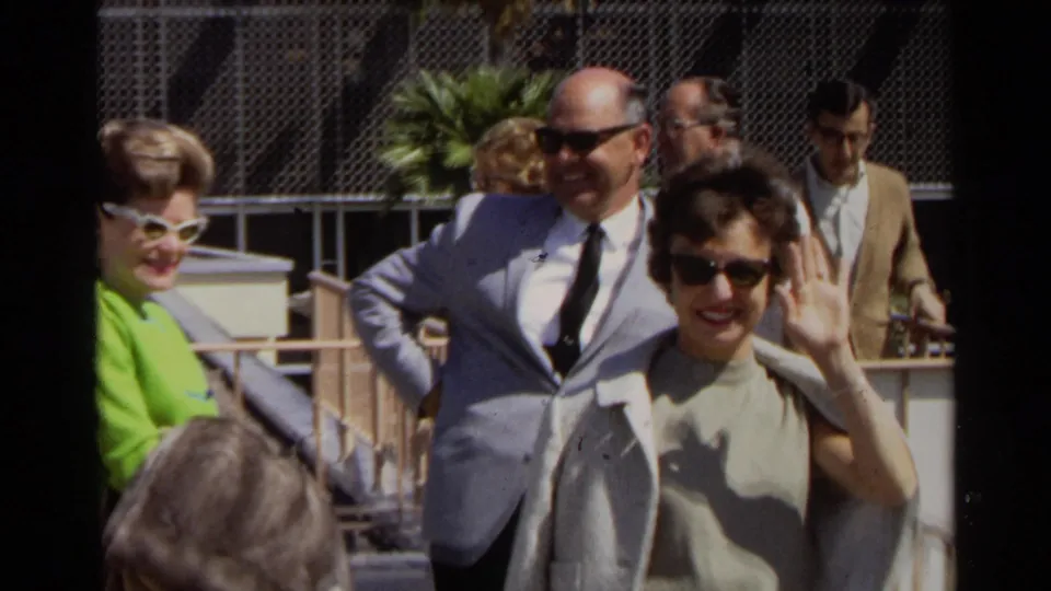 A woman in beige waves toward the camera as formally dressed adults gather on a sunny Scottsdale walkway in the 1968 home-movie clip.
