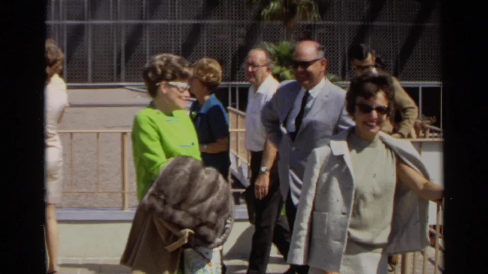 Adults in dress clothing walk past the camera at the Scottsdale event, with the lime green outfit and beige dress prominent in the 8mm frame.