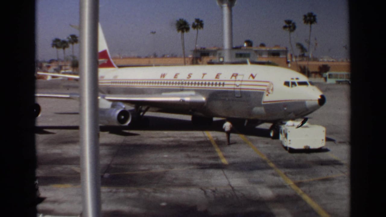 1968 Scottsdale-area home-movie frame showing a Western Airlines jet at the tarmac with ground activity and palm trees beyond.