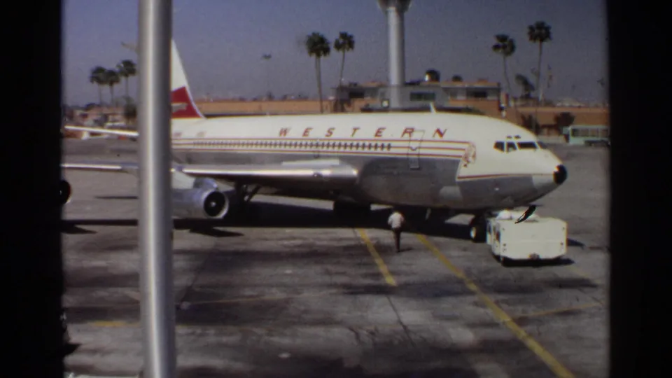 1968 Scottsdale-area home-movie frame showing a Western Airlines jet at the tarmac with ground activity and palm trees beyond.