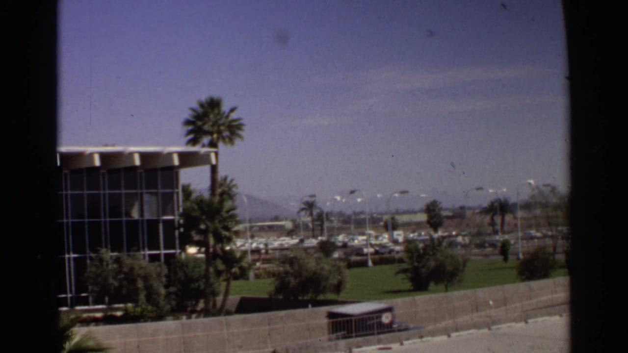 1968 Scottsdale street scene showing a mid-century glass-fronted building, tall palms, green landscaping, parked cars, and distant mountains.