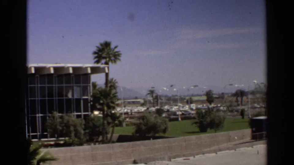 Moving 8mm view in Scottsdale, Arizona, with a glass building, palms, a roadside wall, green lawn, distant cars, and mountain backdrop.