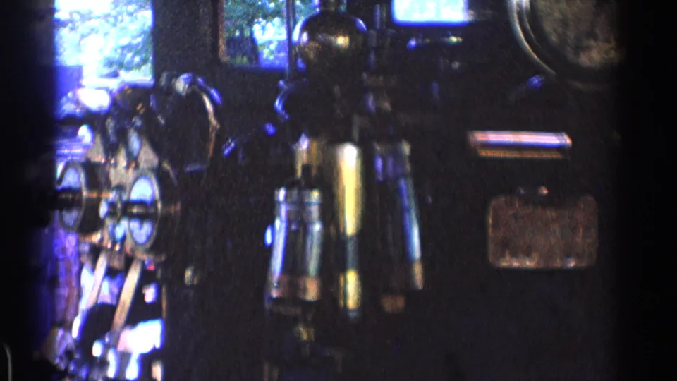 Wider dark cab view with clustered train controls, several windows, analog instruments, and reflective metal handles in archival 8mm footage.