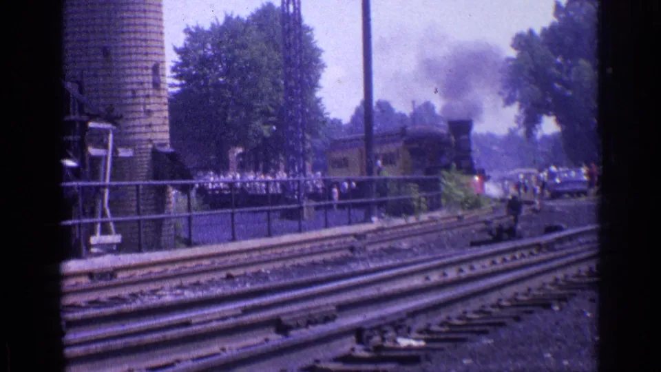 1976 Chicago 8mm frame showing a steam locomotive on multiple tracks, with smoke, a brick silo, trees, and spectators behind a fence.
