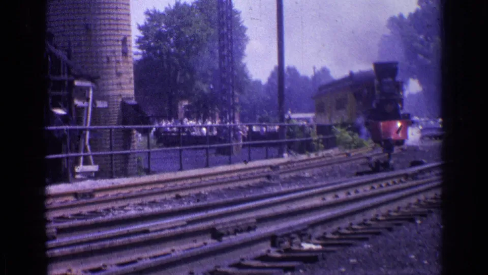 Steam locomotive in a Chicago rail setting, seen near the right edge with foreground tracks, fencing, trees, and distant spectators.