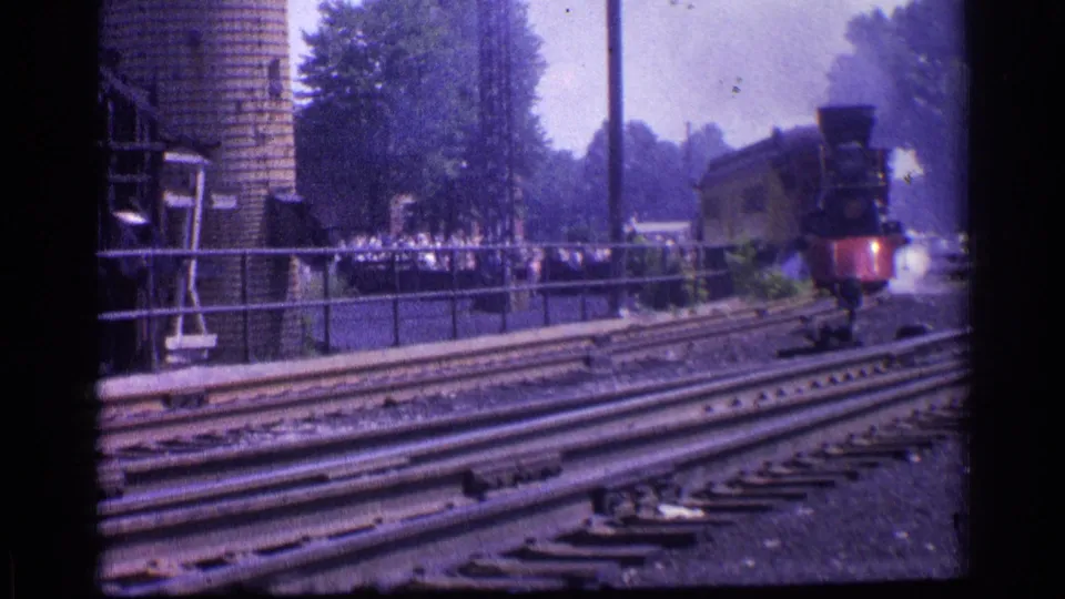 1976 home-movie frame of a steam locomotive moving along Chicago tracks, with smoke at right and a fenced crowd near a brick silo.