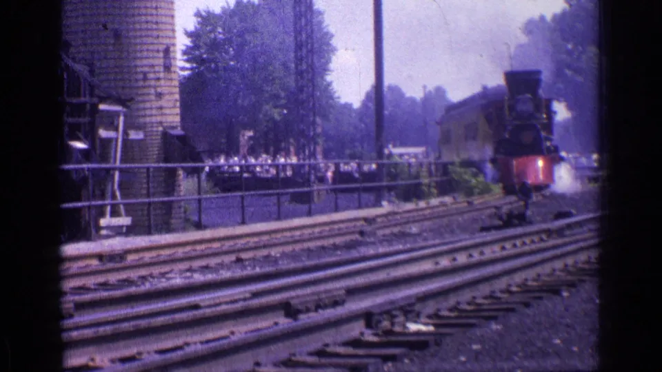 Chicago 1976 8mm frame showing the steam locomotive farther along the rails, with smoke, multiple tracks, trees, and spectators behind fencing.