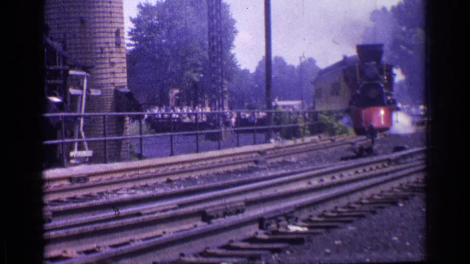 In 1976 Chicago home-movie footage, a black steam locomotive with a red cowcatcher approaches from the distance past rails, a brick silo, trees, and small trackside figures.