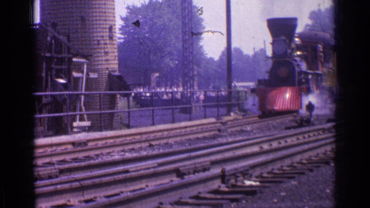 Steam locomotive draws closer beside station tracks and a brick silo.