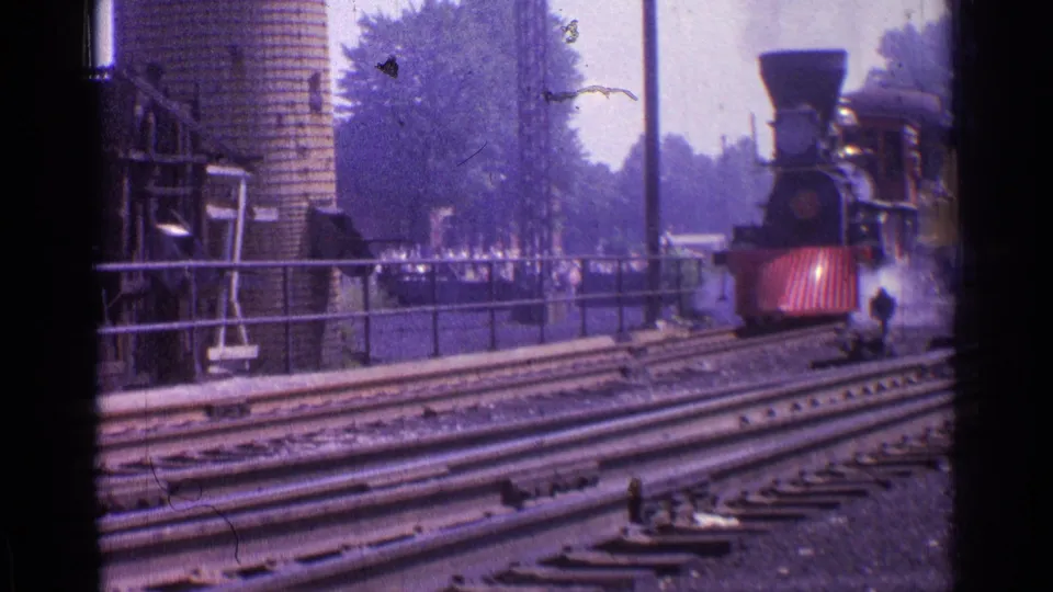 The locomotive moves closer in the 1976 Chicago scene, with smoke around the engine, red front panels, rails in the foreground, and small people visible near the platform area.