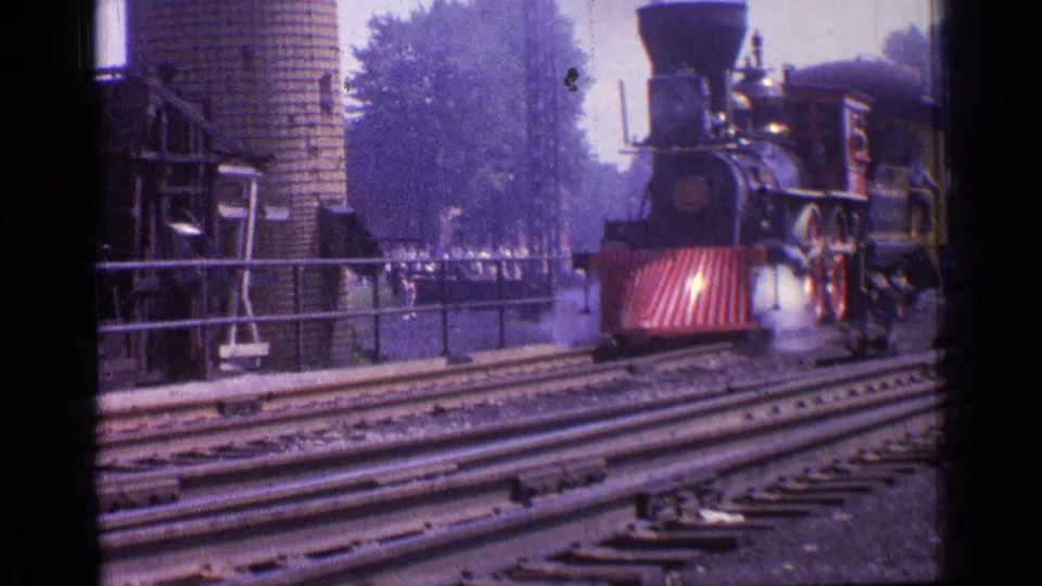 The black steam locomotive advances into a closer view, showing the red cowcatcher, round headlamp area, side rods, smoke near the wheels, station rails, and a brick silo behind it.