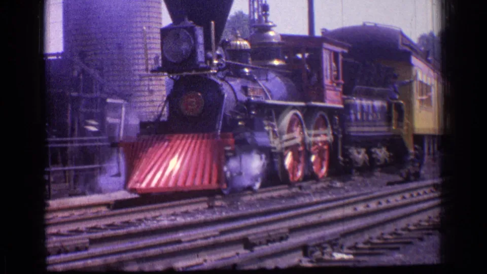 At the end of the 1976 Chicago clip, the locomotive fills the frame with red wheels, a red cowcatcher, visible mechanical rods, smoke at track level, and passenger cars trailing behind.