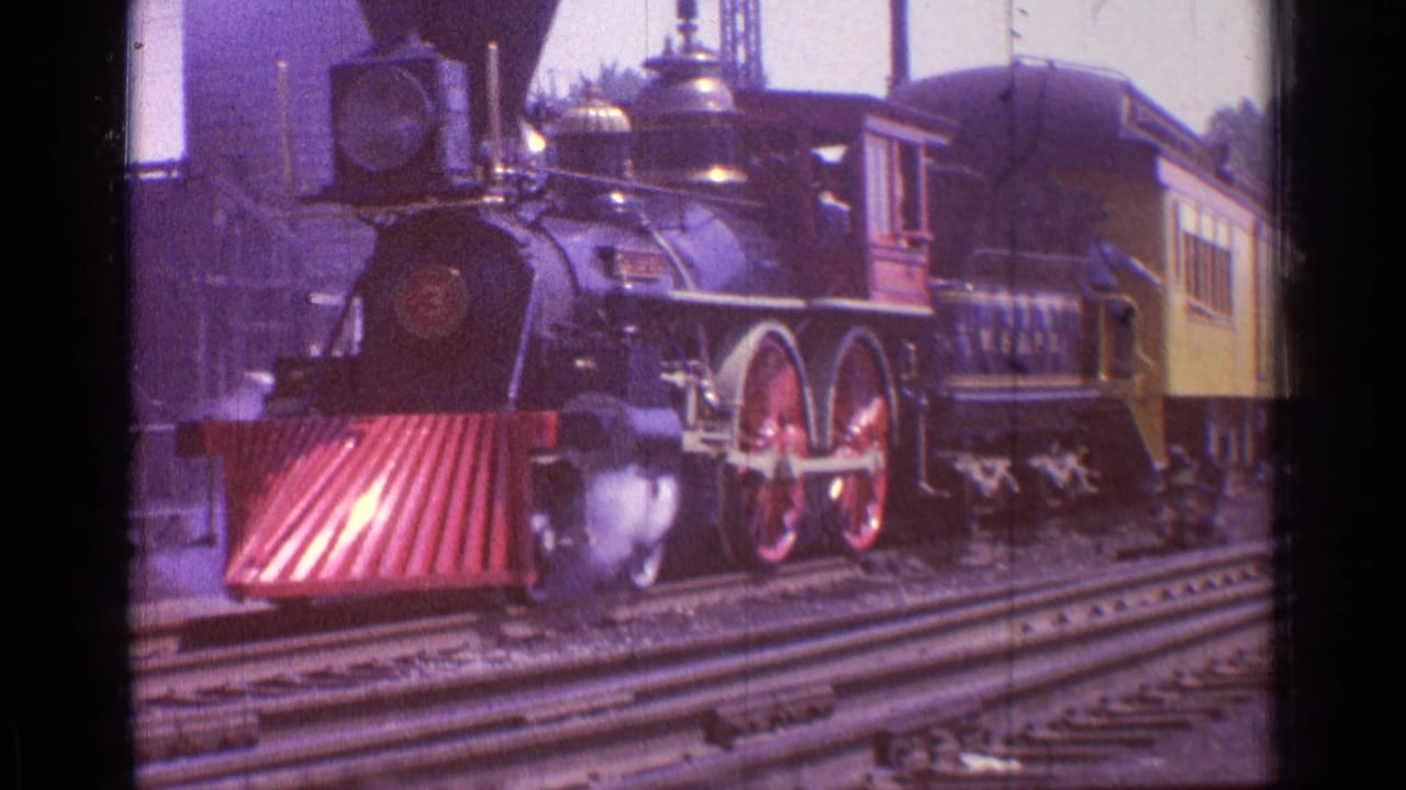 Black steam locomotive with red wheels and red cowcatcher beside a yellow passenger car.