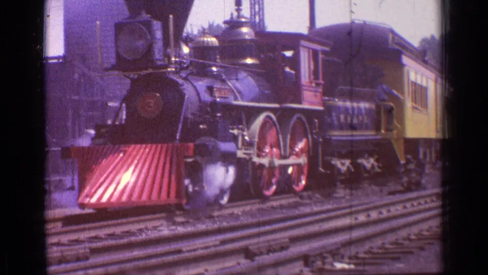 1976 Chicago 8mm frame showing a black steam locomotive with red wheels, smoke and a yellow passenger car on rail yard tracks.