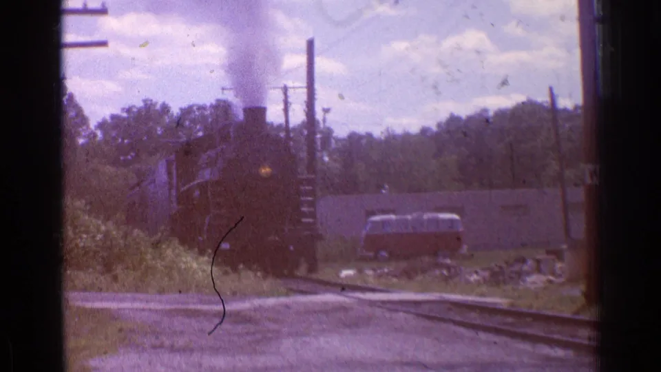 1976 Chicago 8mm frame showing a steam locomotive near a crossing, with smoke, utility poles, rails and a light-colored vehicle in the background.