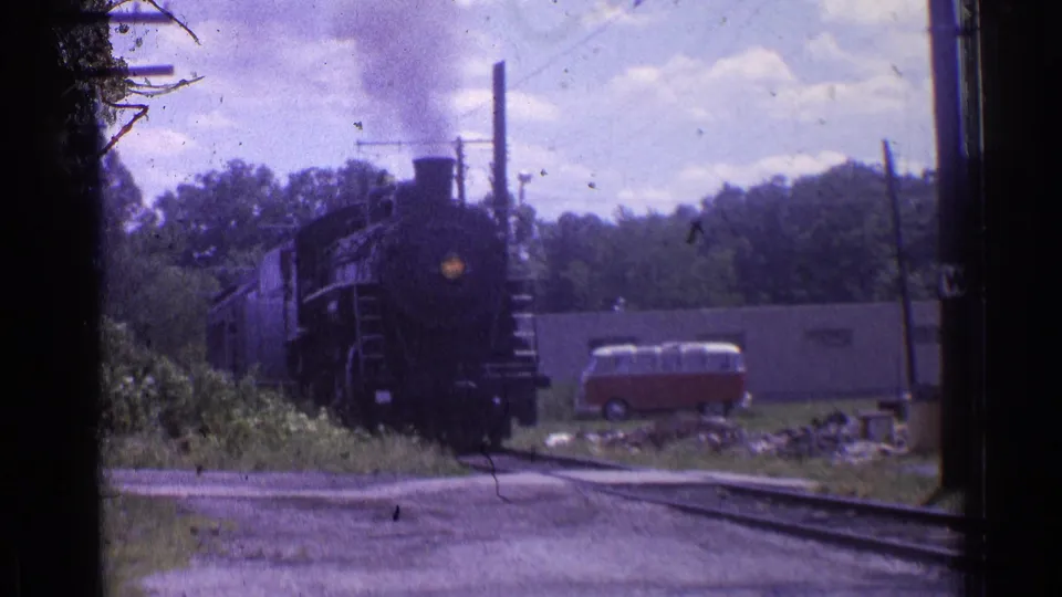 1976 Chicago-area 8mm frame of a black steam locomotive approaching on tracks with trees, poles, a small building, and a red-and-white van nearby.
