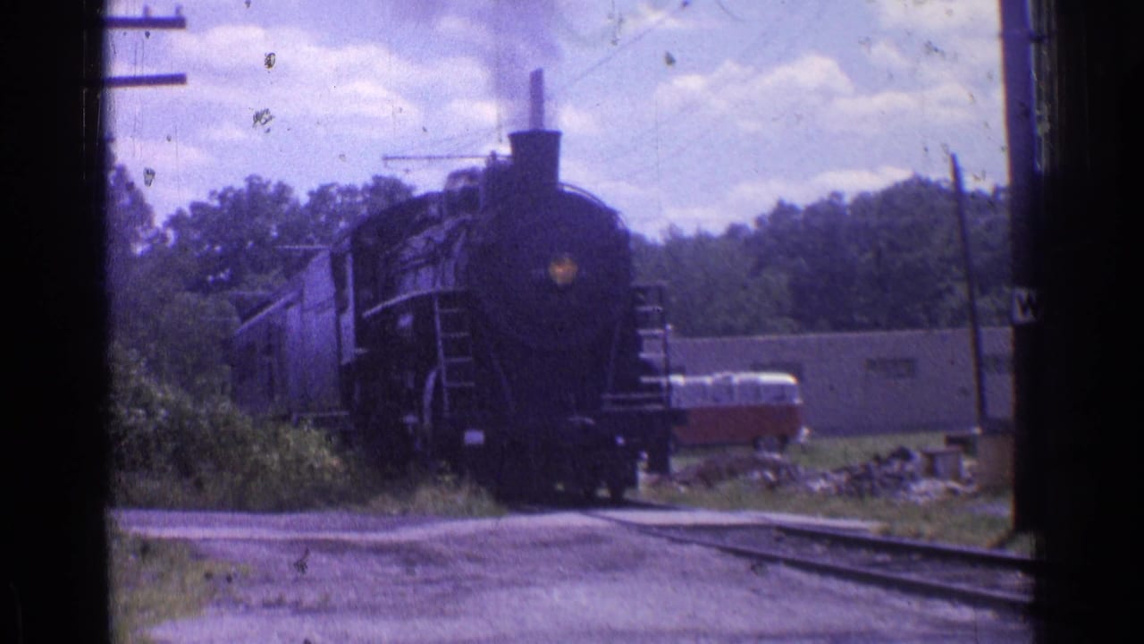 Steam locomotive draws closer on rails with passenger cars behind and a red-and-white van near a low building.