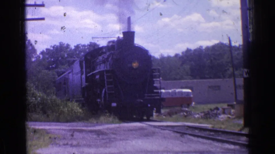 1976 Chicago-area 8mm frame showing the steam locomotive closer to camera, with passenger cars trailing and a van parked near a low building.