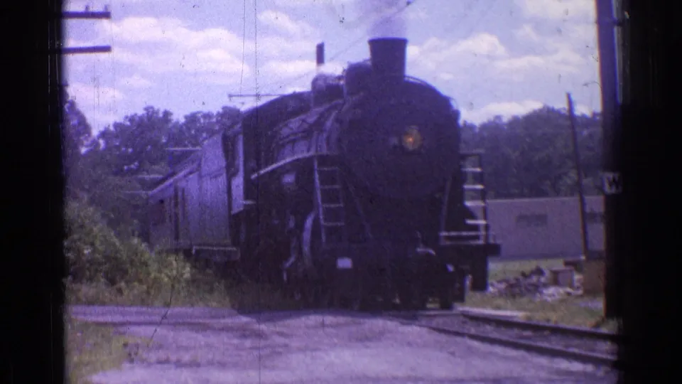 1976 Chicago-area 8mm frame with the front of a black steam locomotive nearing the camera, passenger cars trailing along the trackside brush.
