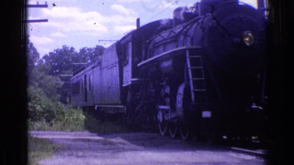 1976 Chicago-area 8mm frame of the black steam locomotive passing close by, with side details, wheels, headlight, and passenger cars visible.