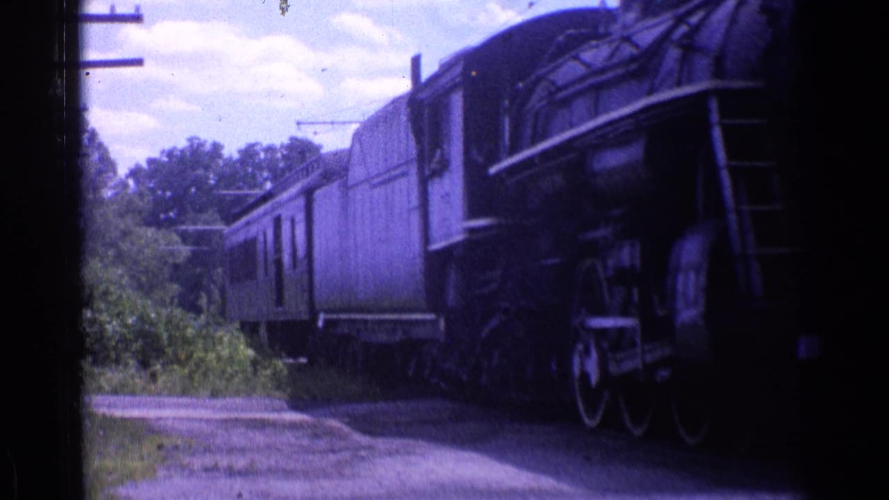 Steam locomotive entering the frame beside passenger cars on a rural track.