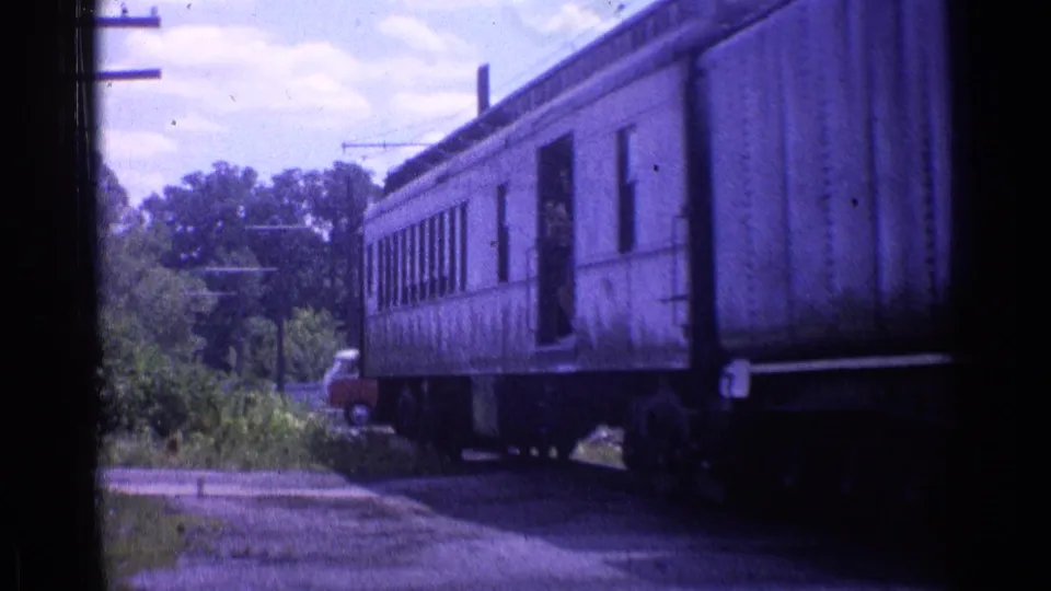 1976 8mm view of a passenger car crossing a small road near Chicago, with brush and poles beside the track.