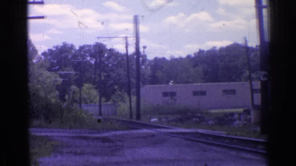 Chicago-area 1976 rail view showing curving tracks, utility poles, dense trees, brush, and a low building under a bright sky.