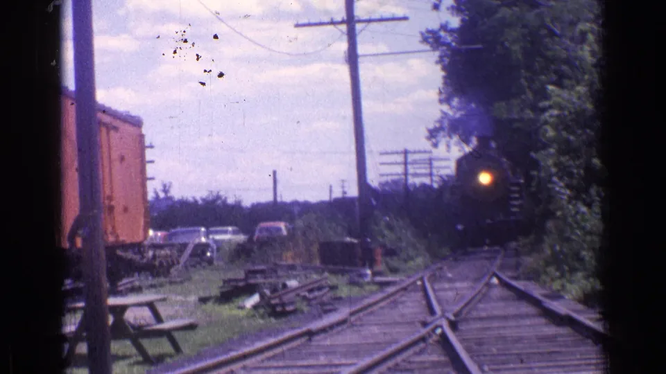 1976 8mm Chicago-area footage shows a steam locomotive approaching with its headlight on, near a red freight car, picnic tables, cars, and trees.