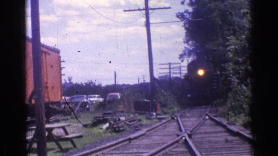 Chicago-area 1976 home-movie frame of a steam locomotive rounding the tracks with a bright headlight, trees, utility poles, parked cars, and a red freight car.