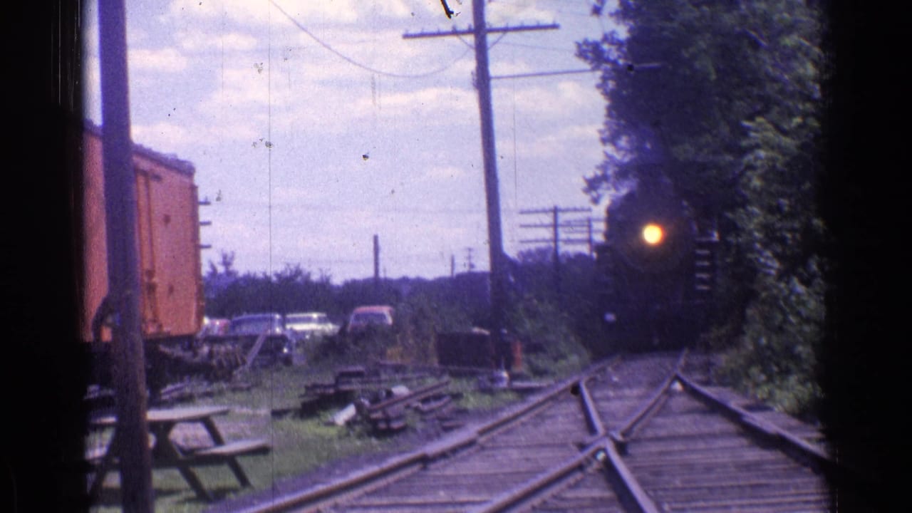 1976 Chicago-area 8mm frame showing a distant black steam locomotive approaching along rural tracks beside a red freight car.