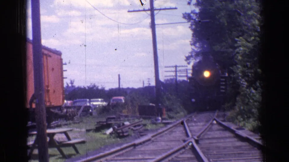 1976 Chicago-area 8mm frame showing a distant black steam locomotive approaching along rural tracks beside a red freight car.