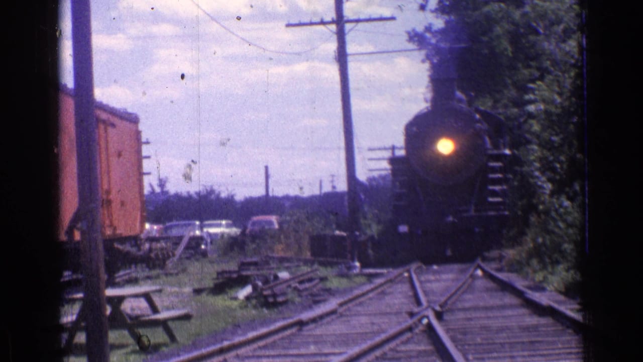 Steam locomotive closer to the camera with its headlight glowing on the track.