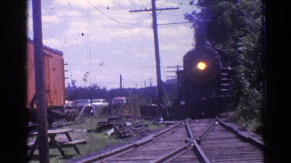 1976 Chicago-area 8mm frame with a black steam locomotive moving toward the camera past grass, trees, utility poles, and parked cars.