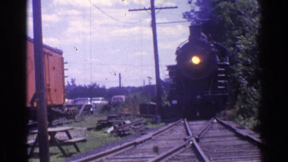 1976 Chicago-area 8mm frame showing the steam locomotive nearing the foreground, with freight car, utility poles, grass, and cars behind it.
