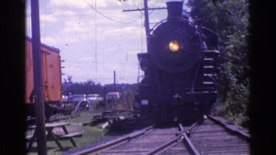 1976 Chicago-area 8mm frame with the steam locomotive close to the camera, headlight visible, framed by tracks, trees, and utility poles.