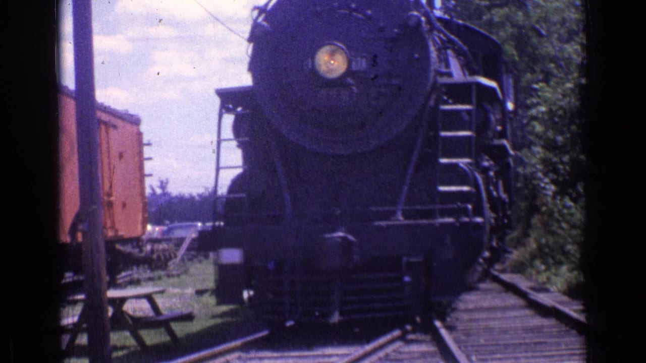 Front of a steam locomotive coming closer on tracks, with trees and rail equipment around it.
