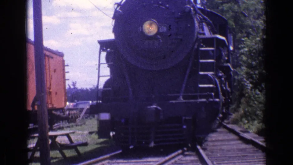 1976 Chicago-area home-movie frame of a dark steam locomotive approaching the camera on tracks near trees and a red rail car.