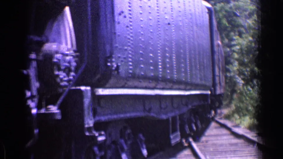 Close trackside view of the riveted tender and lower wheels in 1970s 8mm footage, with foliage lining the rail corridor.
