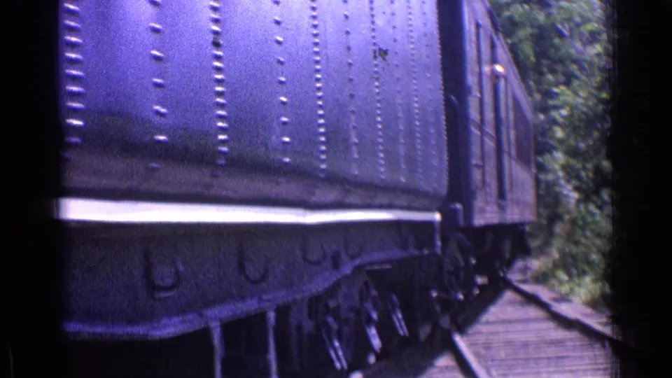 The steam train tender moves away along wooded rails in a 1976 Chicago-area home-movie clip, with purple color shift and grain visible.